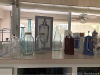Full view of glass jars, beverage dispenser box image, blue French press, brown flask, tall blue-tinted glass bottle, and other glass jars on shelf