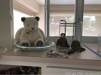Wide view showing a white plush bear sitting inside a clear glass bowl next to two small glass head dolls and a chrome metal rack.