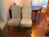 Two wood-framed high back cushioned chairs, upholstered in beige fabric with vertical oval pattern, showing overall front view in a room with wood floor.