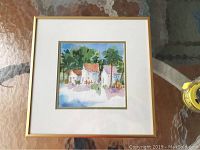 Framed watercolor painting showing three white houses with reddish roofs, trees and foliage in background, matted and framed in gold-tone frame.