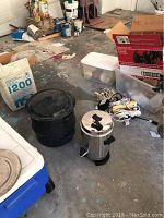 Photo showing a black enamel speckled kettle, stainless steel percolator coffee maker, power cords, and other miscellaneous items on a floor.