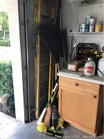 Garage corner with 8ft yellow metal ladder, black plastic rakes, brooms and mops with wooden handles leaning against the wall near a countertop with small bottles.