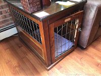 Full view of wood dog crate end table showing glass top, wood and metal grate sides, and front door with latch. Visible chewing damage on wood frame edges.
