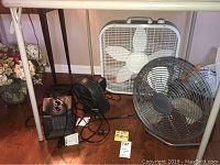 Lasko floor fan, smaller black round table fan, 17-inch round table fan, Holmes ceramic heaters grouped under a table on wooden floor.