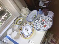 Various Mikasa Meadow Sweet dishes, ceramic platters with colorful patterns, small vases, and crocks on a tabletop by window natural light.