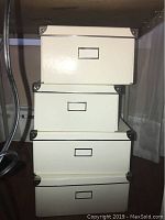 Four stacked cream colored cardboard storage boxes with metal reinforced corners and front label holders.