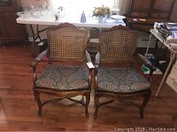 Pair of caned seating wooden armchairs with floral patterned cushions on hardwood floor.