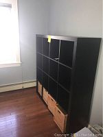 Front angle view of black IKEA cubby shelving unit with four wicker baskets in bottom compartments, showing the shelving unit against the wall on a hardwood floor.