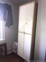 Storage cabinet in a room corner near window with 4 closed white doors framed in beige laminate.