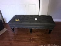 Front view of dark gray rectangular ottoman bench with button tufting and nail head trim on the edges, standing on hardwood floor.