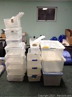 Stack of about 20 clear and blue plastic storage totes of varying sizes in basement room.