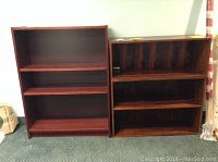Two wood bookshelves side by side; one is a three-shelf reddish wood bookshelf, the other is two-shelf darker wood bookshelf with wood grain finish.