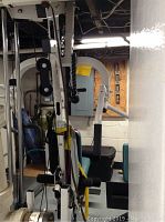 Side view of the white metal frame home gym with black padded seat and rope attachment on pulley system underneath basement ceiling.