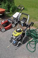 Pressure washer and green garden hose positioned on driveway with lawn mower in background