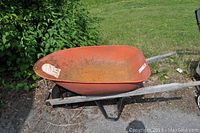 Orange metal wheelbarrow with visible rust and wooden handles and frame on grass surface.
