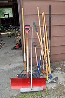 Front view of assorted garden tools leaning against a wall including snow shovels, rakes and a lawn edger.