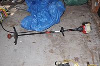 Full length view of the gas-powered weed whacker, showing its straight long shaft, handle grip, engine housing, fuel tank and cutting head with string trimmer inside a garage setting.