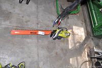 Photo showing the entire Ryobi hedge trimmer with orange blade cover, lying on a concrete floor near some coiled cables and green metal machinery.