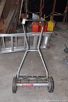 Full view of manual push lawn mower with metal handle in front of ladder and gas cans in background