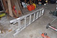 Aluminum extension ladder folded lengthwise resting on a garage floor with background tools and containers.