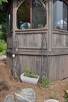Two metal garden trellis frames standing side by side against wooden structure with rectangular plastic plant trough in front with some plants.