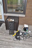 Photo of metallic lantern with geometric patterns, plastic storage box, pond pump with cords, Noma solar spotlight box on wooden deck.