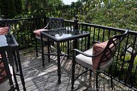 Two tall ornate metal patio chairs with pink cushions and a matching glass-topped square metal table on a wooden deck with outdoor greenery in background.