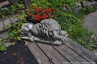 Full side view of the cement lion statue placed on wooden planks with flowers and greenery in the background