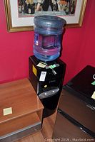 Full view of black classic water cooler with an empty bottle on top in a room with red walls and wooden furniture.