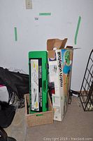 Photo showing boxed volleyball net and badminton net in carrying case placed on the floor leaning against the wall.