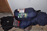 Photo showing two dark-colored sleeping bags rolled in storage sacks, a black cooler bag, and two laptop bags stacked on the floor next to a door.