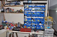 Full view of metal storage rack with many blue bins containing screws, bolts, nuts, and batteries. Workbench below with additional tools and containers.