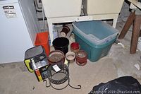 Multiple items on concrete floor near wall including cooler bag, boxed heater, planter pots, storage bins, and metal plant stand.