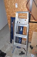 Aluminum step ladder standing upright with blue metal saw horses behind it against a plywood wall.