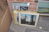 Three framed prints leaning against a brick wall. One is a Tom Thomson poster on board, and two other framed prints with forest and stream landscape images.
