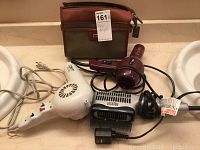 Photo shows three hair dryers on a counter along with a men's toiletry bag; cords are visible and hair dryers are in various colors (white, maroon, black)