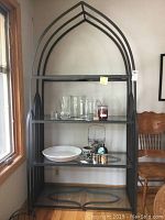 Full view of the black metal framed etagere with glass shelves, showing arch top design and some glassware displayed on shelves.