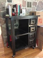 Full view of black wooden kitchen island showing five small drawers on the right, two open shelves in the middle, and cookbooks, radio, and candle holder on top surface