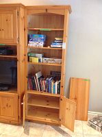 Front view of the pine shelving unit showing multiple shelves with various books and games on them, two enclosed cabinet doors at the bottom, adjacent to a similarly finished piece of furniture and loose wood panels.