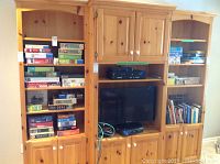 Wide view of wooden shelving unit filled with puzzles, board games and books across multiple shelves with TV multimedia area in center.