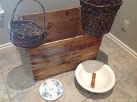Full view of all items: large wooden crate, two woven baskets hanging and positioned, ceramic wash basin with embedded thermometer, and ceramic lid with floral decor on floor.