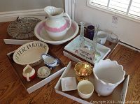 Wide view of collection showing antique ceramic wash basin with matching pitcher featuring pink band, clear glass divided serving dish, oval butter pudding dish, small pitchers, and other ceramic and glass items arranged on floor in cardboard trays.