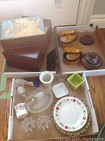 Photo showing wooden jewelry boxes with decorative lids, several smaller wooden and ceramic containers, glass dishes, and candle holders in white cardboard trays