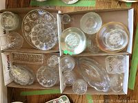 Top-down view of assorted pressed glass platters, plates, bowls, and a sherry glass arranged in boxes on a wooden floor.