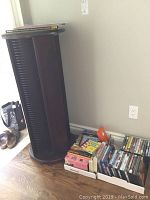 Wooden dark wood tall CD tower standing on floor with two white boxes next to it containing assorted VHS and DVD movies. Vinyl records stacked on top of the tower.
