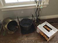 Photo showing brass coal bin with gloves and wooden brush inside, next to black ash pail, fireplace tools stand, and boxed fire logs on wood floor beneath window