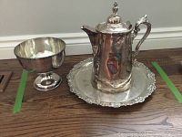 Silver plated thermos jug, chalice, and tray set displayed on wooden floor.