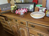 Wooden cabinet with decorative items including framed wall art, ceramic jug, two decorative bowls, and two vintage-style clocks on top.