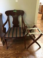 Photo showing the full wood chair with armrests and upholstered seat cushion next to the wooden luggage rack with fabric straps on hardwood floor.
