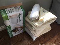 Stacked wool blankets with white bolster pillow on top, next to boxed HoMedics memory foam bed pillow on wooden floor